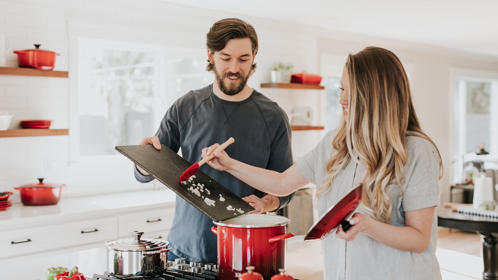 Couple cooking together in a bright white kitchen with open walnut shelving and red cookware